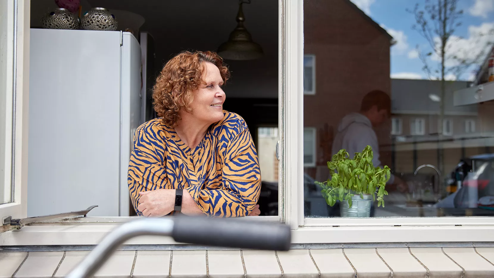Beeld bij bericht over de kostendelersnorm. Een vrouw kijkt uit het keukenraam en je ziet een medebewoner op de achtergrond.