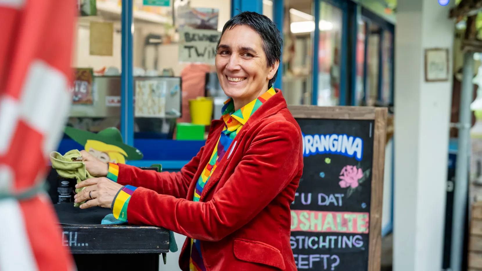 op de foto staat Stephanie Caton, eigenaar van ontmoetingsplek de Boshalte, in een rood pak. Ze staat voor de uitgiftebalie van haar horeca, op het terras.