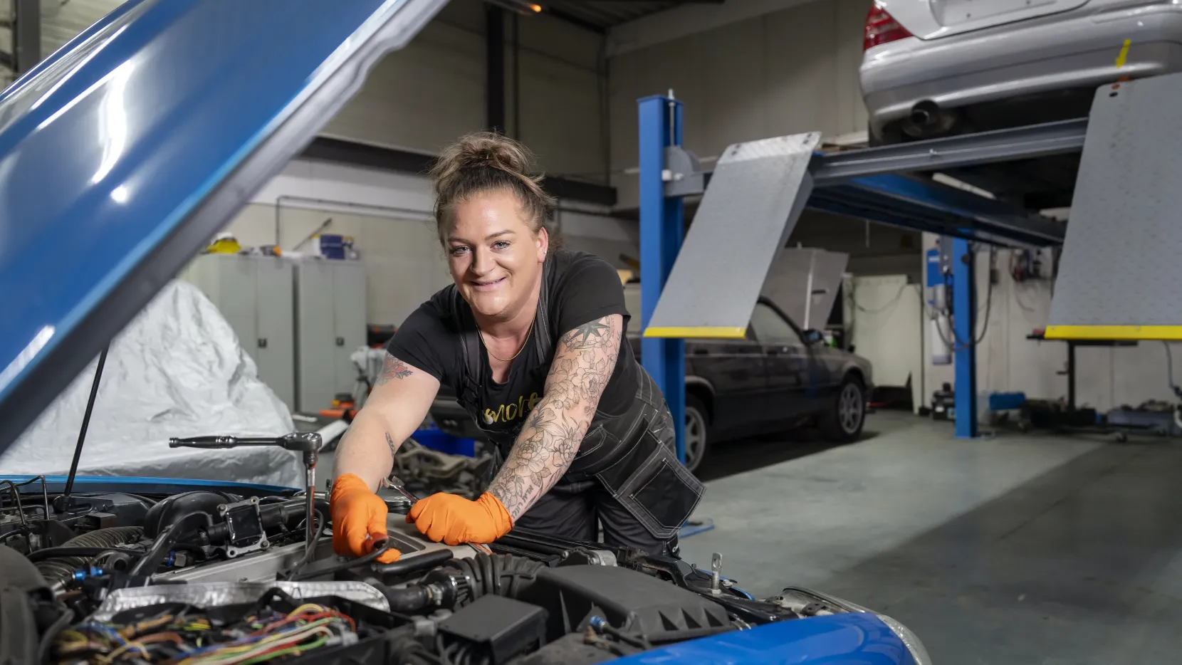 Portret van Jacintha. Ze is aan het werk onder de motorkap en lacht naar de camera. Ze heeft een werkoverall aan en oranje werkhandschoenen.