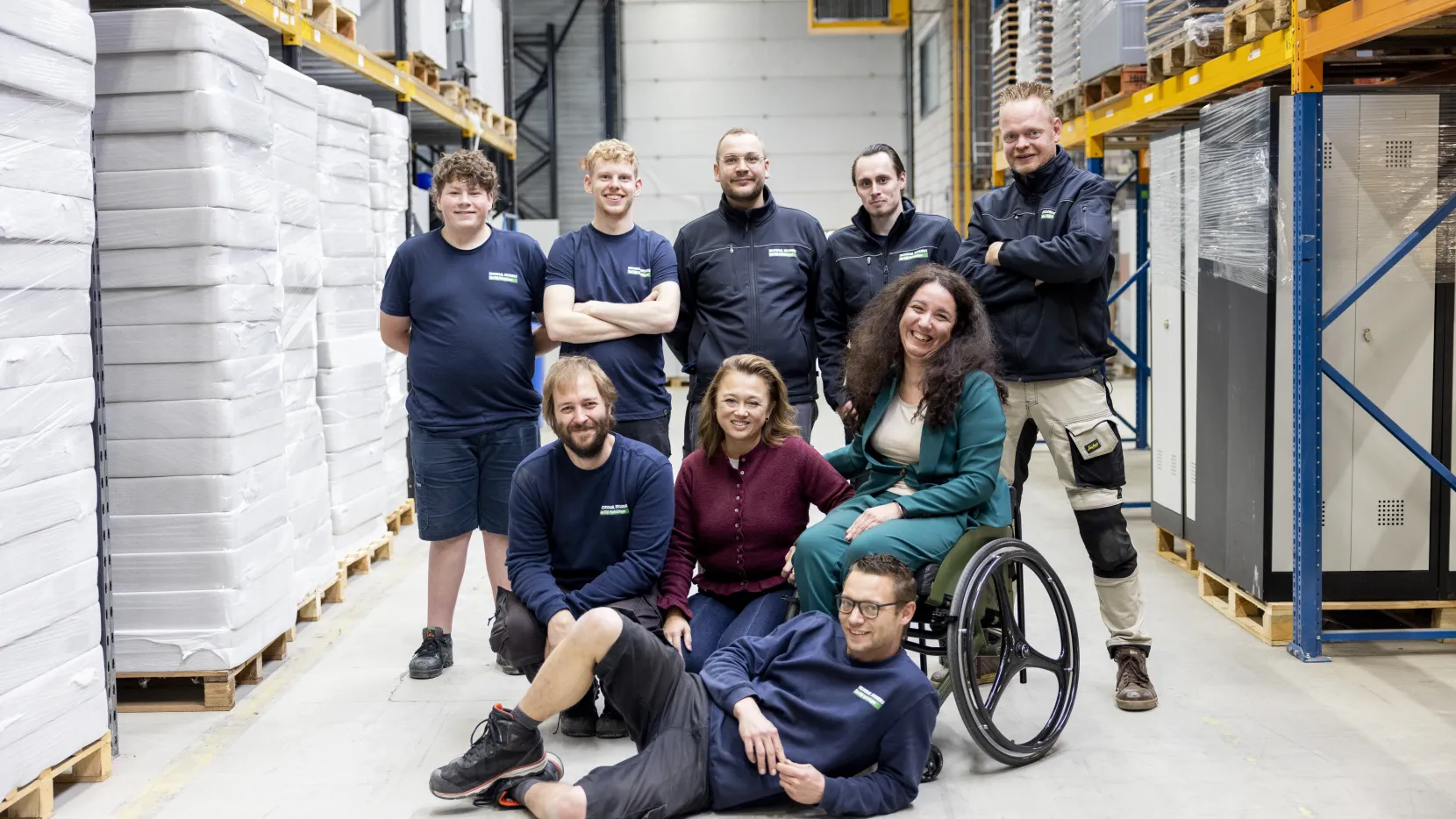 Portret van Nadia. Ze poseert samen met een aantal van haar collega's van Breedweer voor de foto's. Ze zijn in een magazijn omringd met grote witte pallets. Ze kijken lachend naar de camera. 