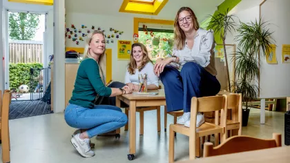 Suzanne van Sinne zit op tafel. Haar nieuwe personeelslid Samantha, hurkt links. Achter de tafel zit een collega van Samantha. ze zitten in een zaal van de kinderopvangopvang met kindertekeningen