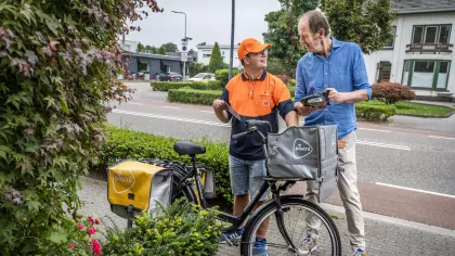 Portret van Daniel Claessen, die ene fiets aan de hand houdt en Willo van Bommel. Ze staan op straat en kijken elkaar lachend aan.