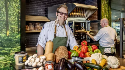 Portret van Wesley Mons die met een voorleesbril in de keuken werkt. Hij staat achter een tafel met groenten. Op de achtegrond zien we de werkgever op de rug.