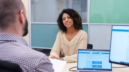 Een gesprek bij UWV. Een vrouw zit aan een bureau. We zien de man met wie zij een gesprek heeft van achteren, zijn achterhoofd en schouder.