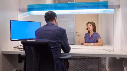 Een man en een vrouw zitten tegenover elkaar aan een bureau. We zien de man van achteren en de vrouw van voren aan de overkant van de tafel. Een blauw beeldscherm staat links op het bureau.