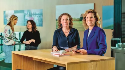 Elles Diepeveen en Damiët Martinot, initiatiefnemers van PGGMama, staan achter een bureau. Op de achtergrond zien we twee vrouwen in met elkaar praten.