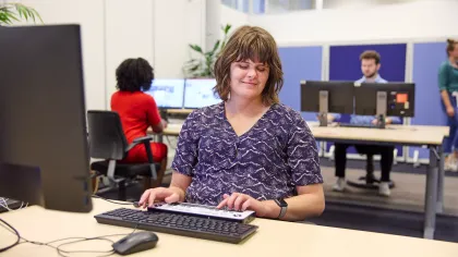 Een vrouw met een visuele beperking zit achter een bureau, dat aangepast is voor haar. Op de achtergrond werkt een dame in het rood en zien we nog twee andere werknemers.