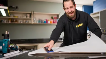 Erik kijkt staat in de keuken en kijkt lachend de camera in. Hij heeft folie in z'n hand om een keuken te gaan wrappen.
