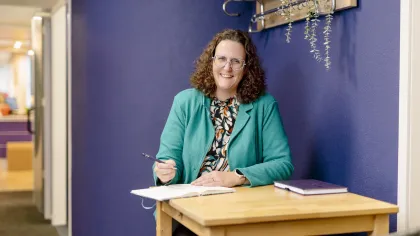 Portret van Nathalie. Ze zit achter een bureau en heeft een pen in haar hand met een kladblok. Ze kijkt lachend de camera in.