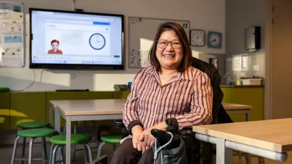 Portret van Joyce. Ze zit in haar rolstoel in haar klaslokaal en lacht naar de camera. Ze heeft een roze rode blouse aan.