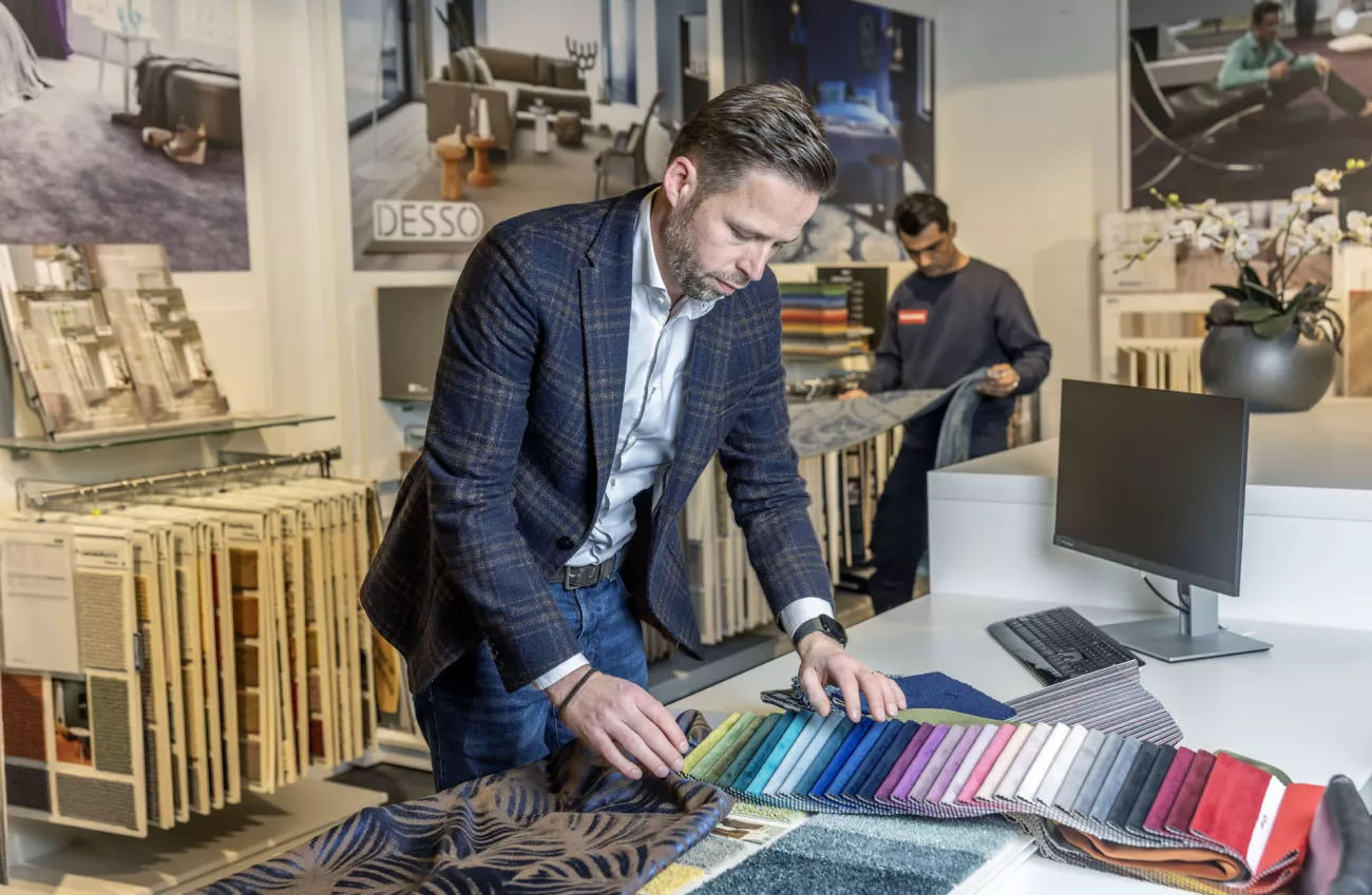 Mathijs Groen kijkt naar verschillende stalen in zijn winkel. Achter hem staat zijn nieuwe werknemer Hamid.