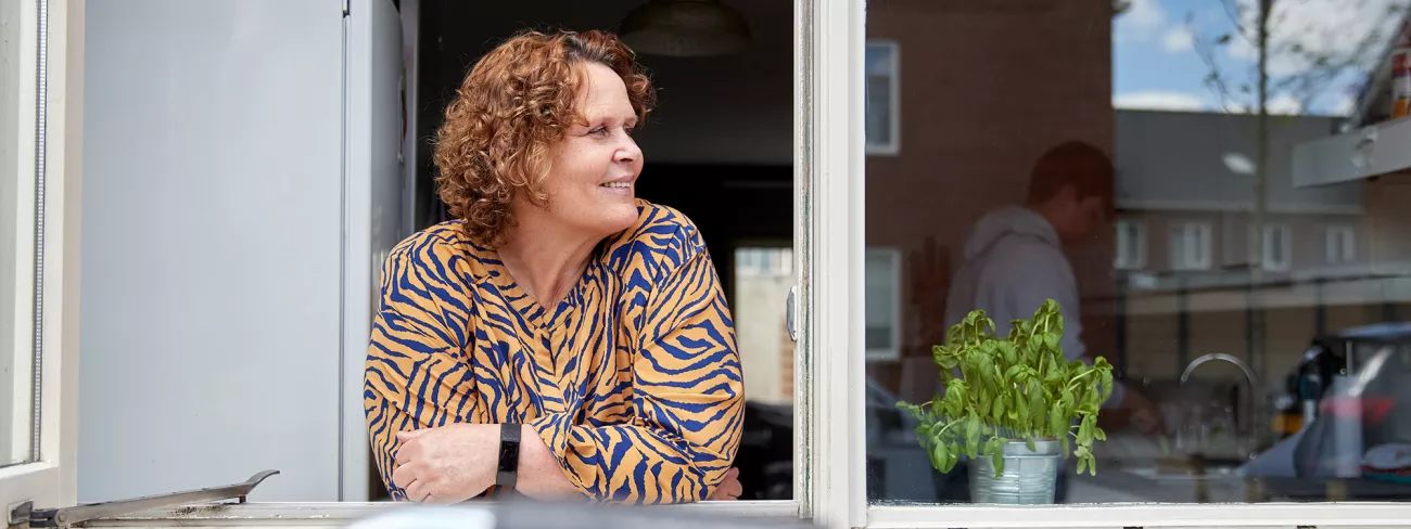 Beeld bij bericht over de kostendelersnorm. Een vrouw kijkt uit het keukenraam en je ziet een medebewoner op de achtergrond.