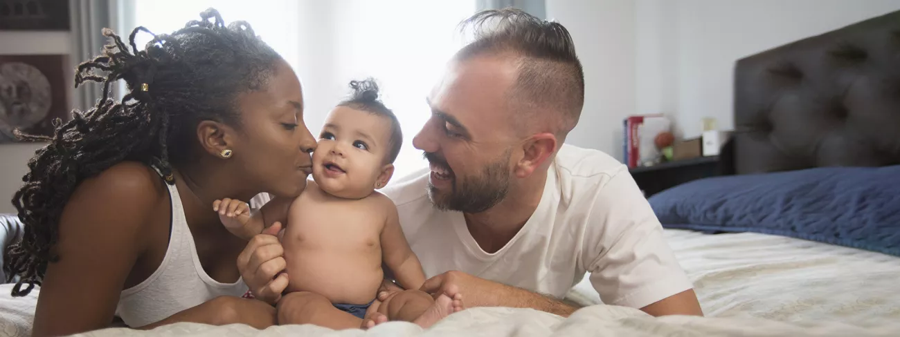 WIEG: Een man en een vrouw liggen op bed. Ze kijken beiden naar de baby die tussen hun in zit. De vrouw kust de baby. UWV / Getty images