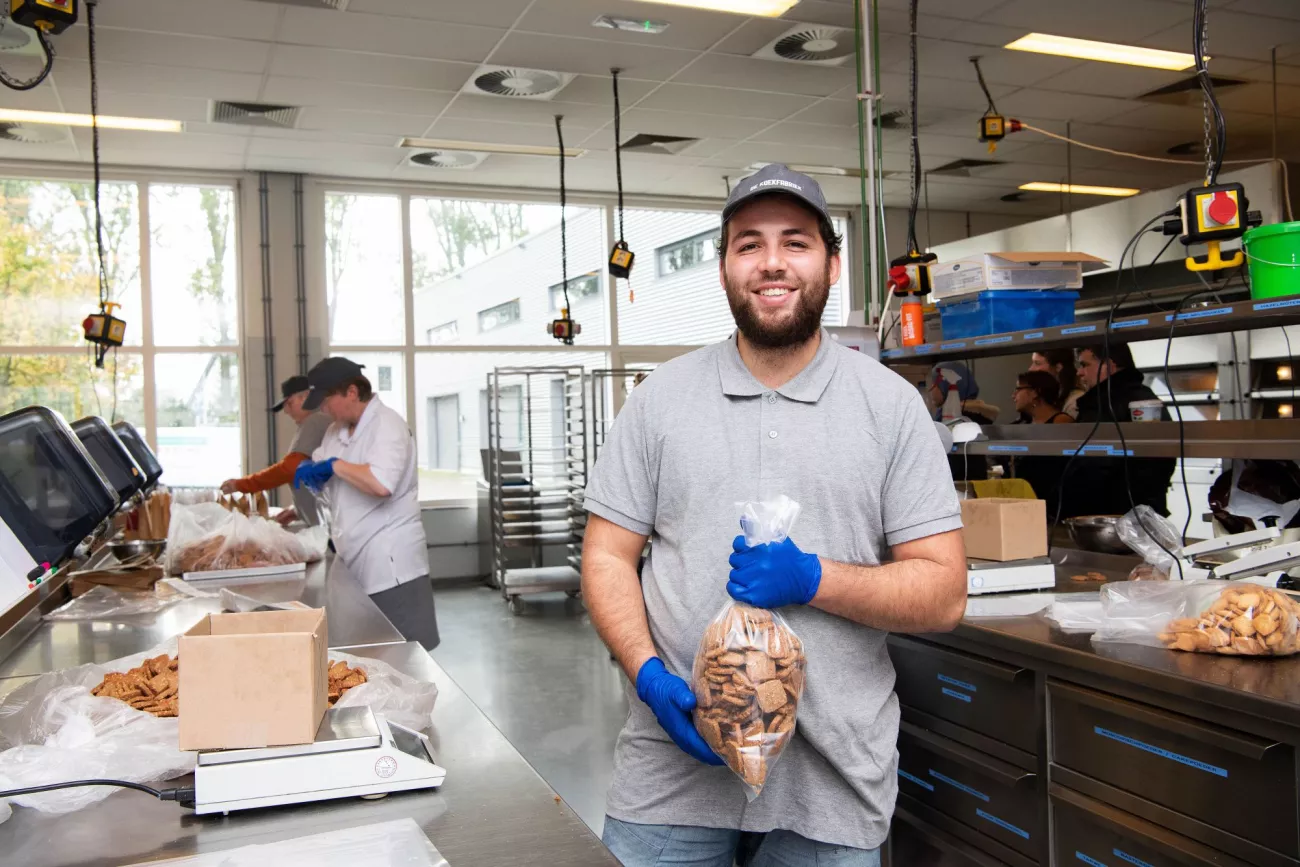 Medewerker van een koekjesfabriek staat trots met een zak koekjes. Op de achtergrond zien we 2 andere collega's werken in de fabriek. UWV - Medea Huisman