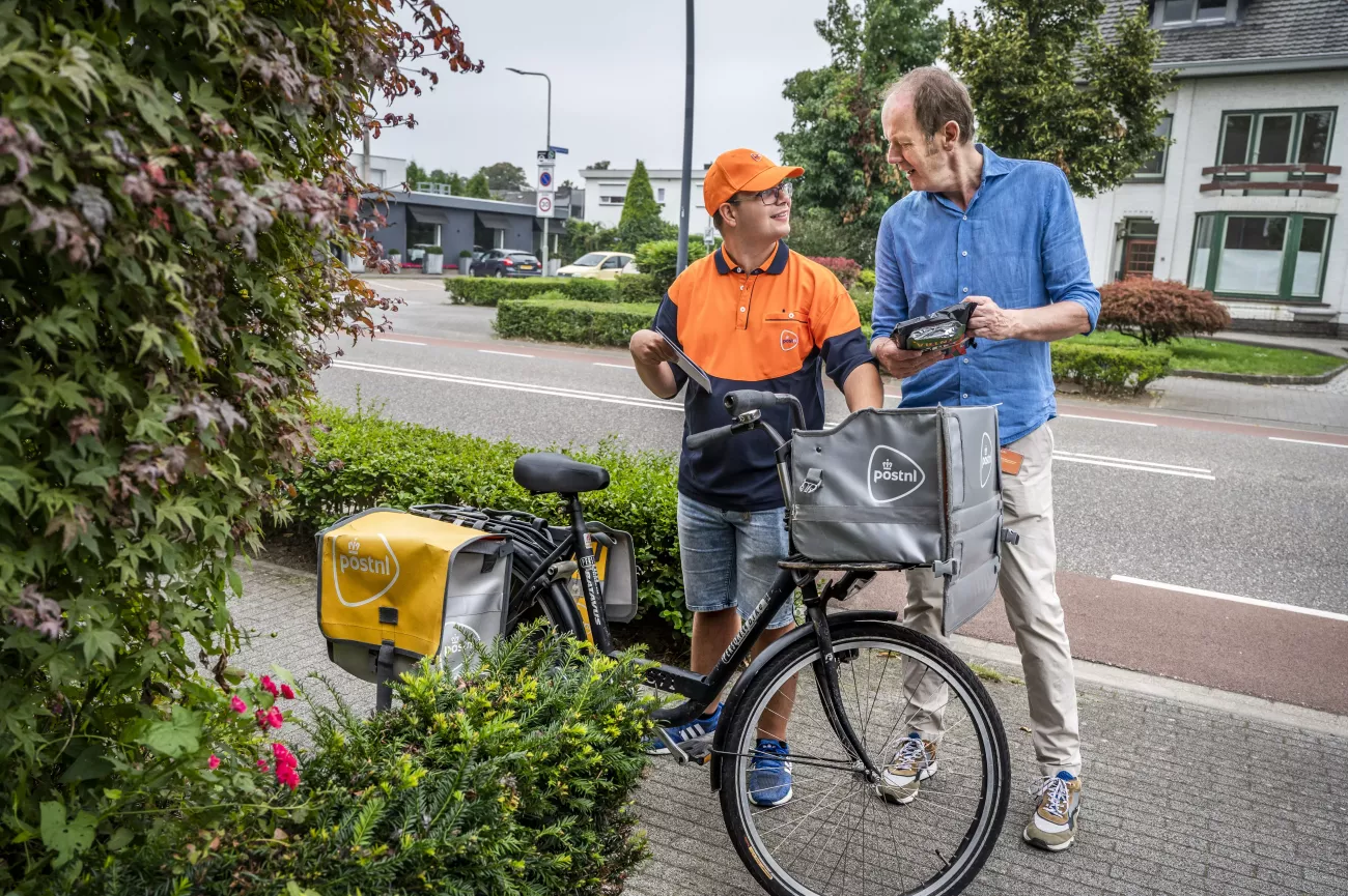 Portret van Daniel Claessen, die ene fiets aan de hand houdt en Willo van Bommel. Ze staan op straat en kijken elkaar lachend aan.