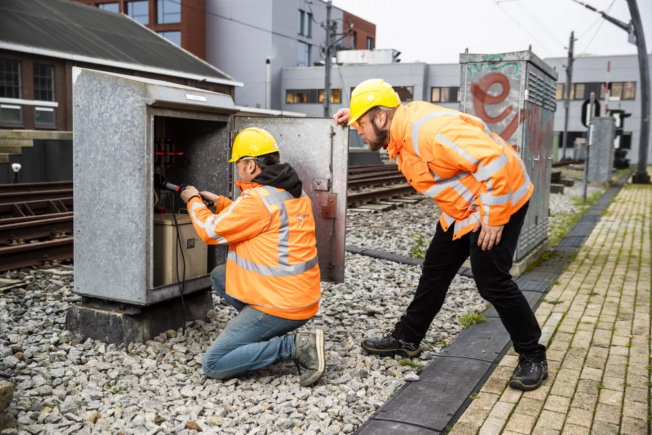 Een railtechnicus in opleiding kijkt in een kastje. Zijn begeleider kijkt toe over de schouder. Ze dragen beiden veiligheidshesjes en helmen.