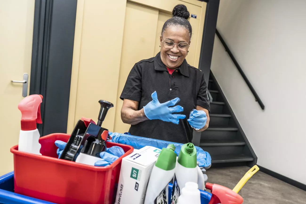 Carmen trekt een handschoen uit achter een kar met schoonmaak artikelen, en lacht