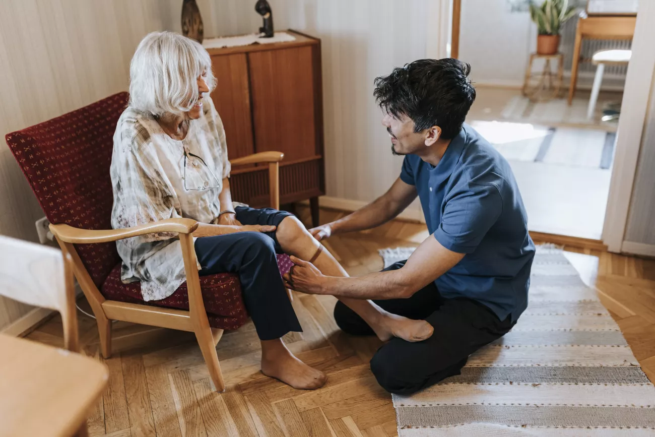 Een zorgverlener helpt een oudere dame in een stoel haar kousen aan te trekken in een huiskameromgeving.