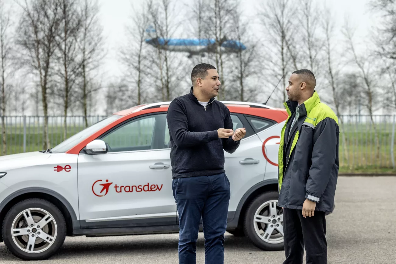 Dave van Amsterdam met medewerker Jardel staan bij een auto van Transdev met op de achtergrond een vliegtuig dat landt.