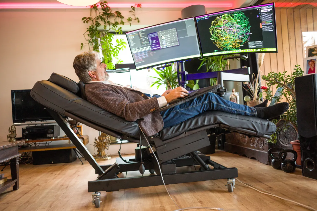 Bjorn ligt in zijn ligstoel en is aan het werk. Hij kijkt naar de 2 computerschermen voor hem en heeft een cursor in zijn hand. 