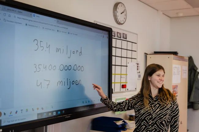Kim staat vooraan in het klaslokaal bij het digitale schoolbord. Ze wijst met haar rechtervinger naar het bord en kijkt de klas.