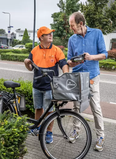 Portret van Daniel Claessen, die ene fiets aan de hand houdt en Willo van Bommel. Ze staan op straat en kijken elkaar lachend aan.