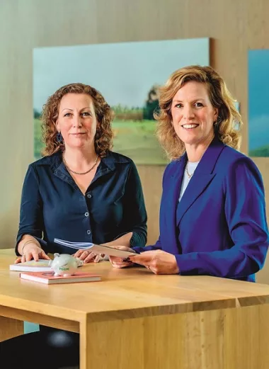 Elles Diepeveen en Damiët Martinot, initiatiefnemers van PGGMama, staan achter een bureau. Op de achtergrond zien we twee vrouwen in met elkaar praten.