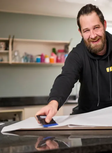 Erik kijkt staat in de keuken en kijkt lachend de camera in. Hij heeft folie in z'n hand om een keuken te gaan wrappen.
