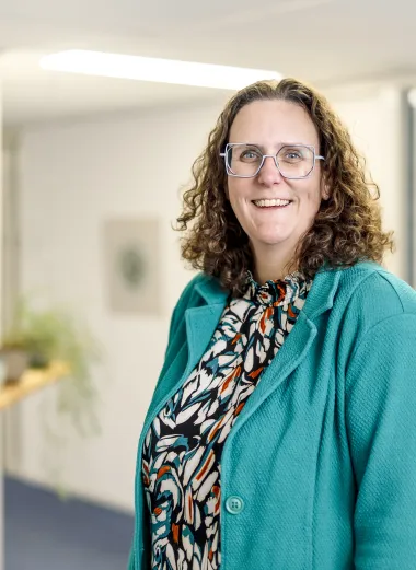 Portret van Nathalie. Ze staat in de gang van haar kantoor en kijkt lachend de camera in.