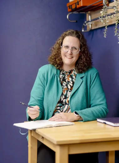 Portret van Nathalie. Ze zit achter een bureau en heeft een pen in haar hand met een kladblok. Ze kijkt lachend de camera in.
