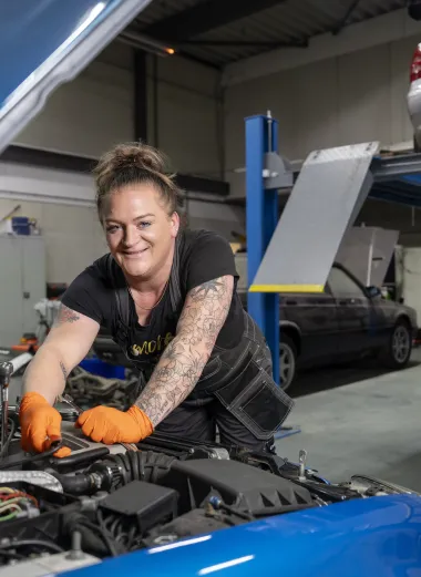 Portret van Jacintha. Ze is aan het werk onder de motorkap en lacht naar de camera. Ze heeft een werkoverall aan en oranje werkhandschoenen.