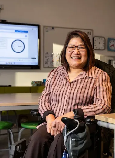Portret van Joyce. Ze zit in haar rolstoel in haar klaslokaal en lacht naar de camera. Ze heeft een roze rode blouse aan.