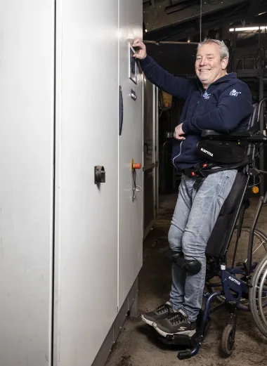 Portret van Ronald. Hij staat in zijn sta-op rolstoel en lacht naar de camera. Hij staat voor een werkkast.