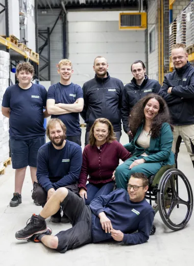 Portret van Nadia. Ze poseert samen met een aantal van haar collega's van Breedweer voor de foto's. Ze zijn in een magazijn omringd met grote witte pallets. Ze kijken lachend naar de camera. 