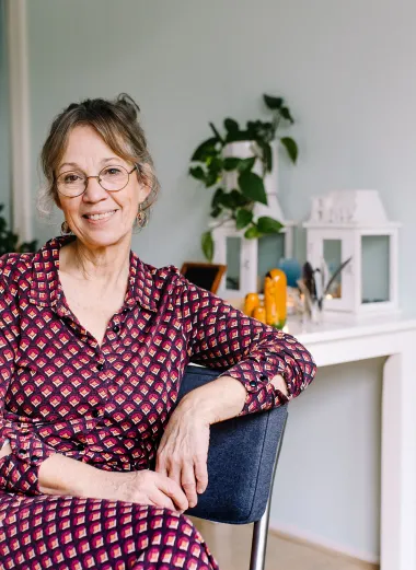 Portret van Conny. Ze heeft een rood roze jurk aan en zit op een stoel. Ze kijkt glimlachend naar de camera. 